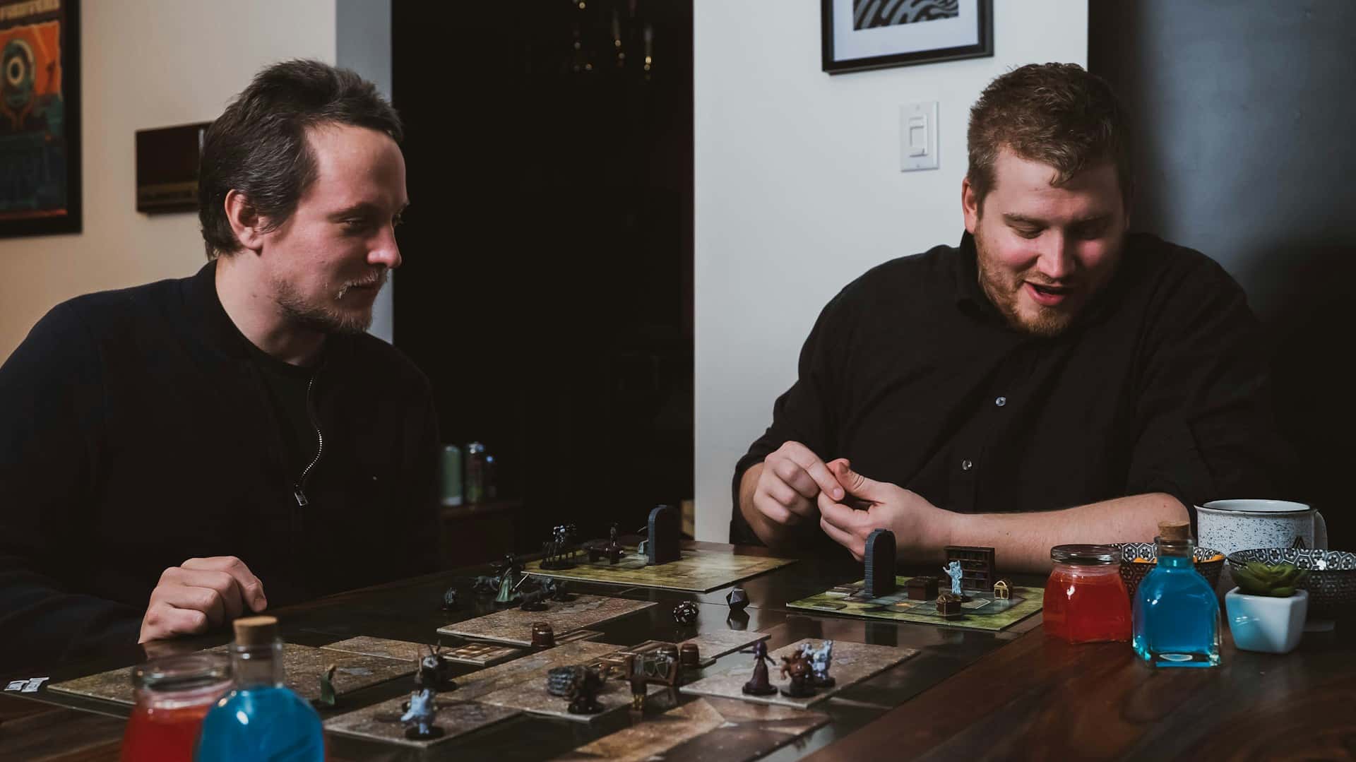 Two players at a tabletop game session with miniatures, dice, and modular map tiles.